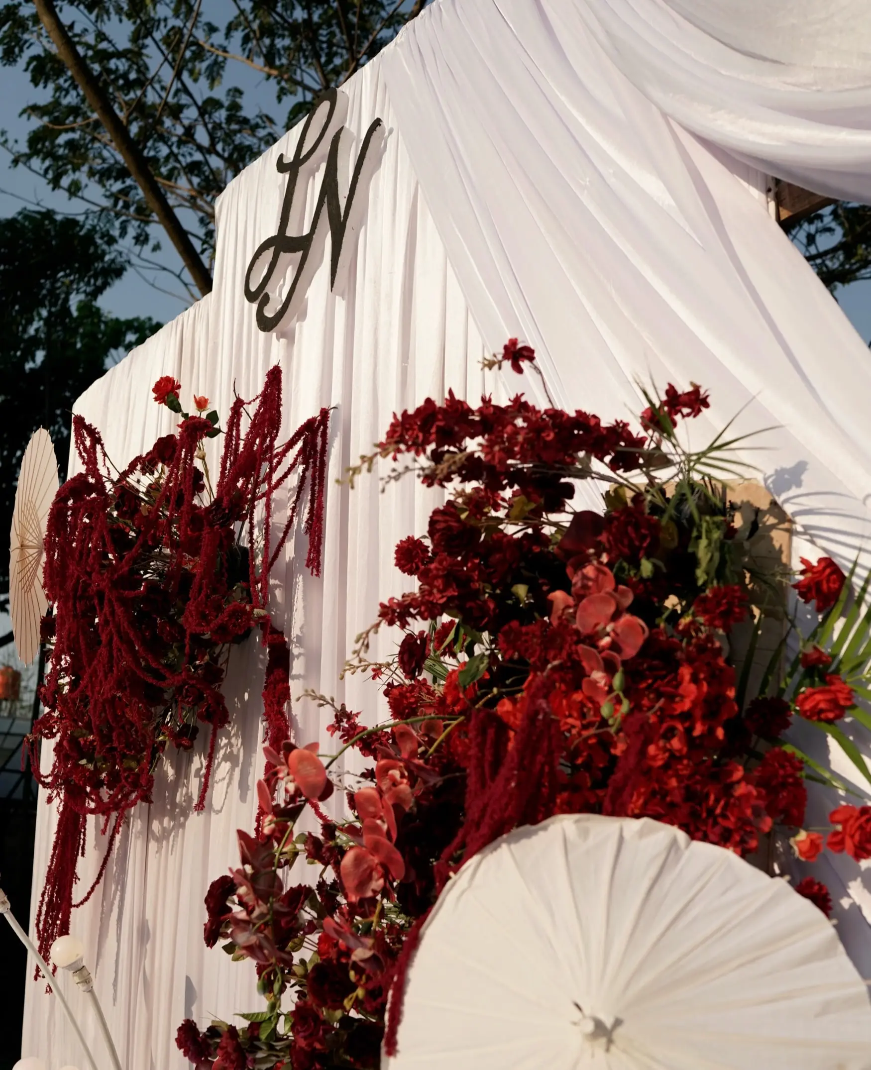 Dekorasi garden party dengan rangkaian bunga merah, kain putih menjuntai, dan payung kertas