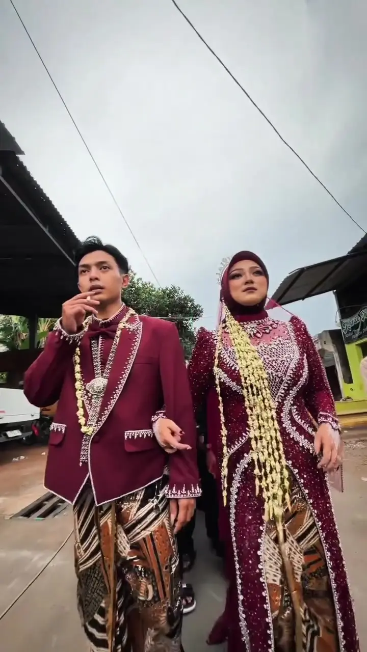 Pasangan pengantin berbusana marun berjalan diiringi keluarga dan tetangga