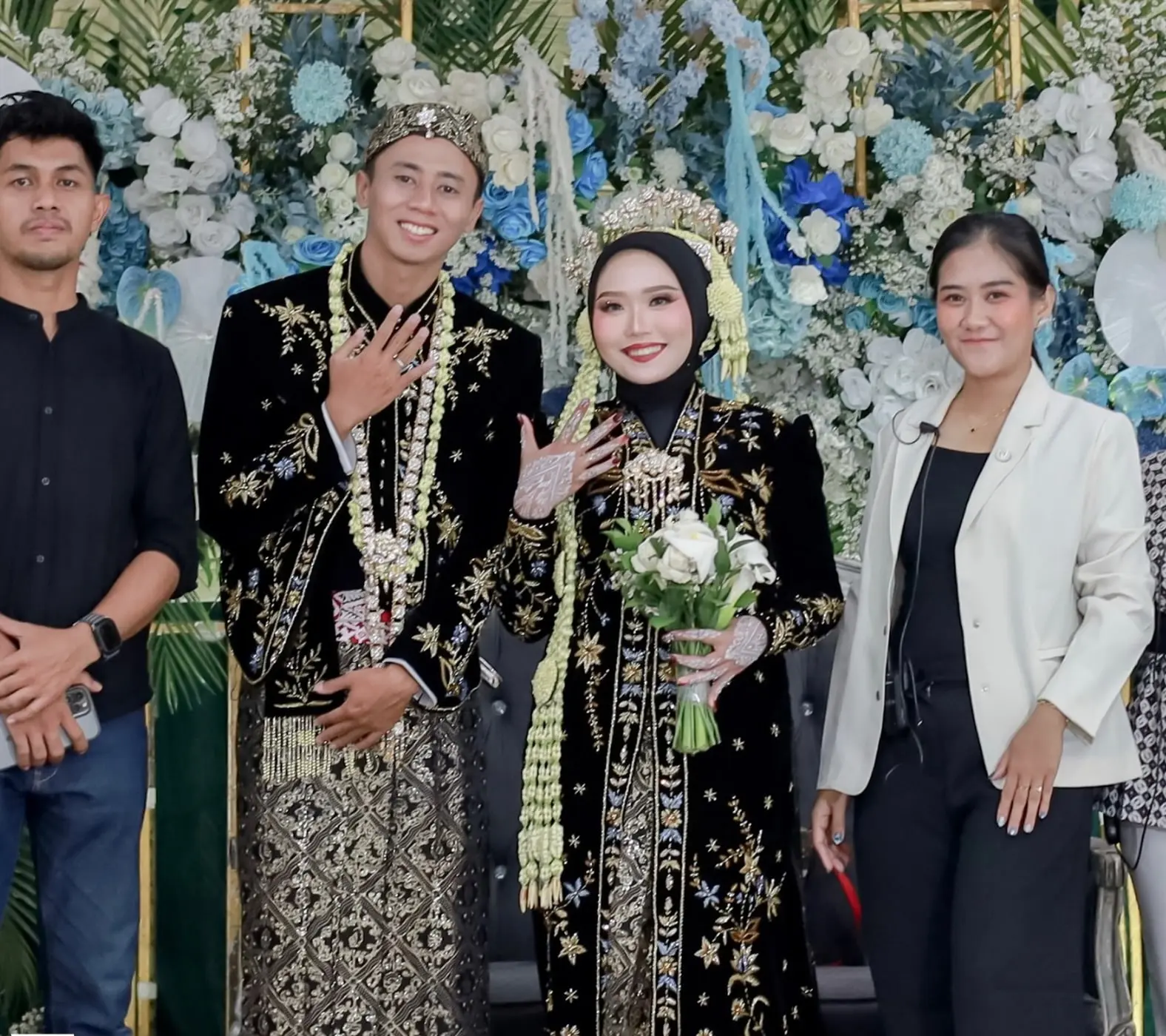Kru JOIEA berfoto bersama pasangan pengantin berbusana Jawa hitam di pelaminan bunga biru