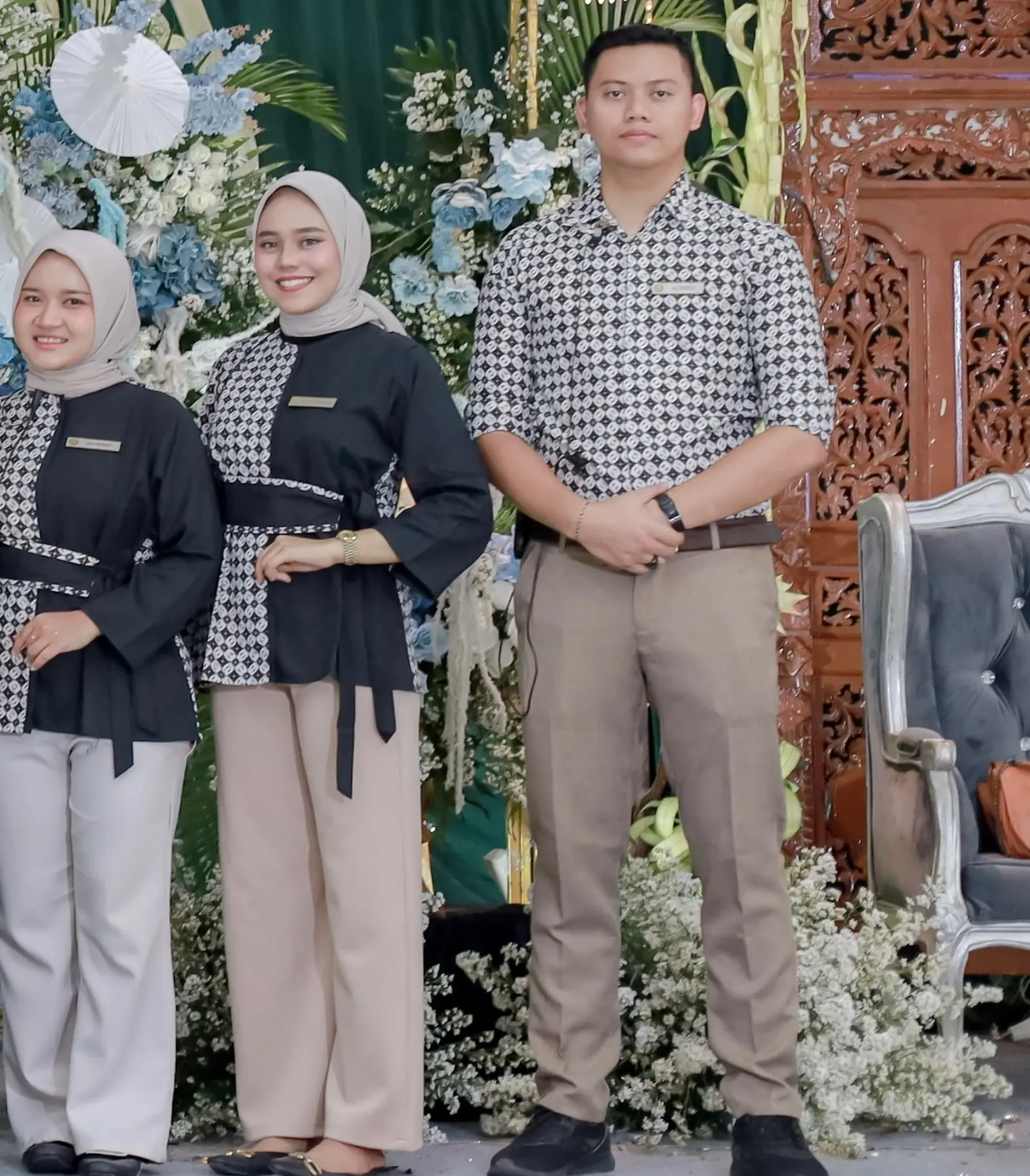 Tiga kru JOIEA berseragam batik hitam putih berdiri di depan dekorasi bunga