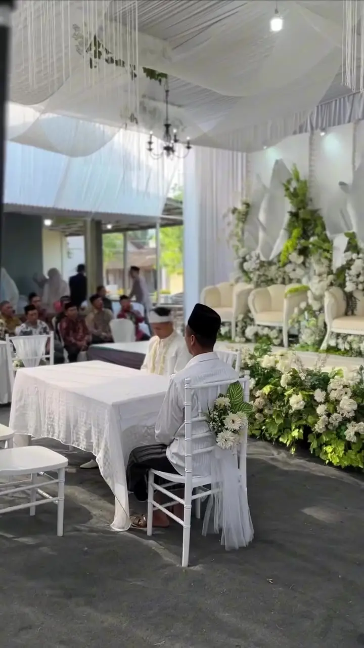 Meja akad bertaplak putih di bawah tenda berplafon kain dengan pelaminan bunga di belakangnya