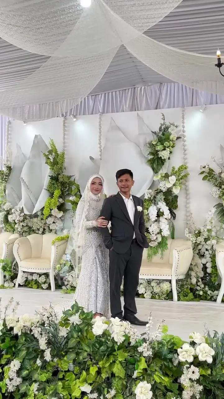 Pasangan pengantin berdiri di pelaminan bunga putih di bawah tenda berplafon kain
