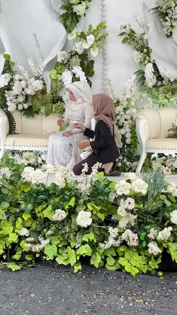 Pelaminan dengan sofa krem, bunga putih, dan dedaunan hijau, pengantin wanita sedang dirapikan riasannya