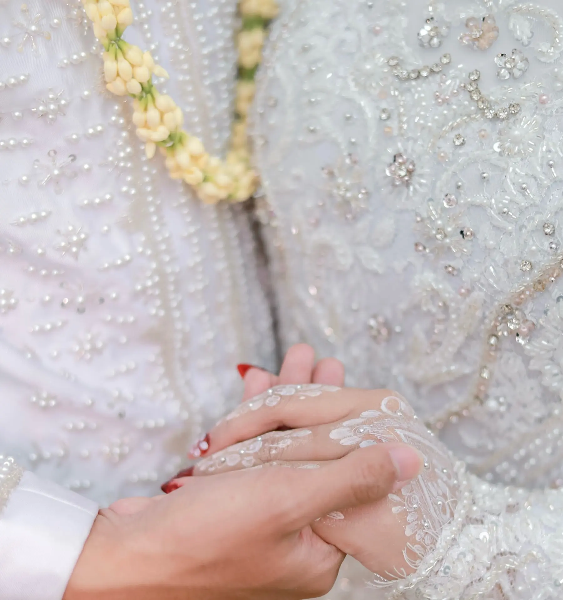 Tangan pengantin saling menggenggam di depan busana putih berpayet dan ronce melati